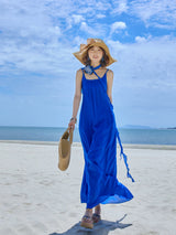 Woman walking on a white beach in a blue low back strap maxi dress, with a straw hat and woven bag