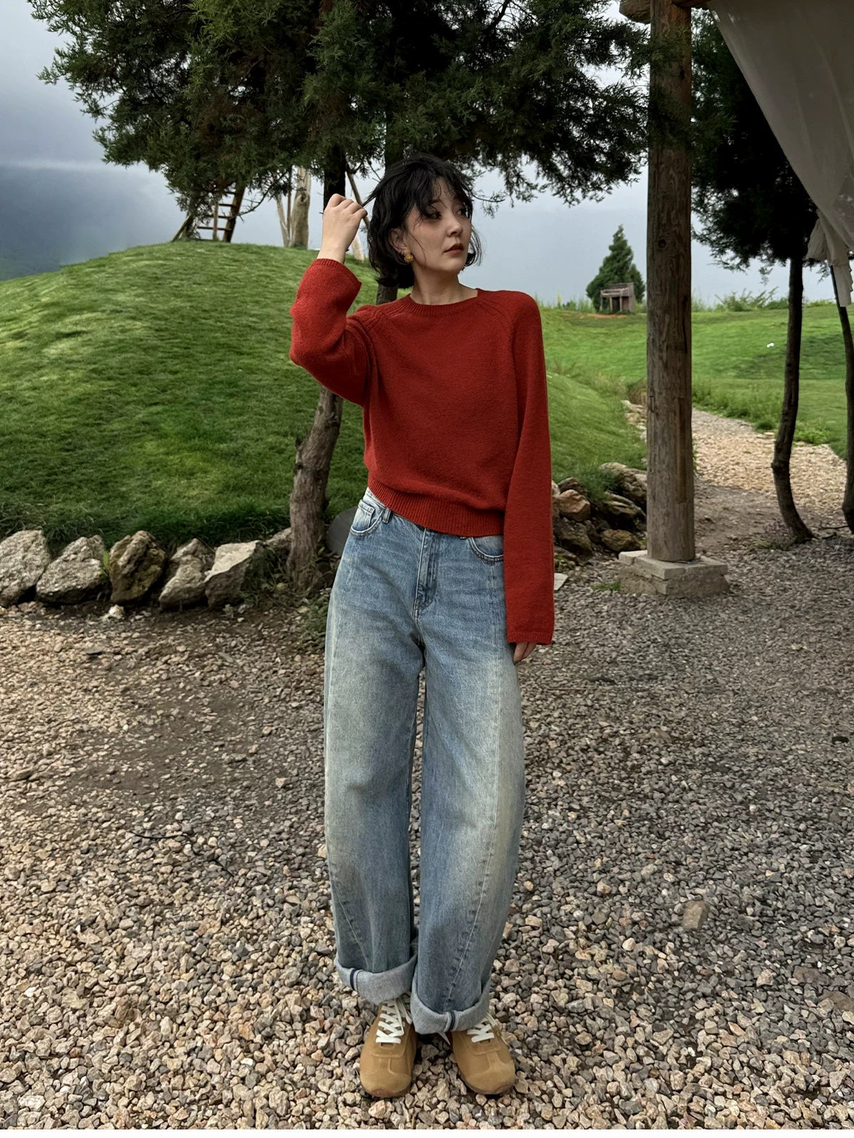 Woman on a gravel path below a grassy hill wearing a red raglan crew sweater and wide jeans