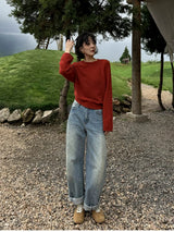 Woman on a gravel path below a grassy hill wearing a red raglan crew sweater and wide jeans