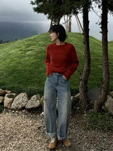 Woman beside pine trees and a grassy mound in a red crew sweater and wide jeans
